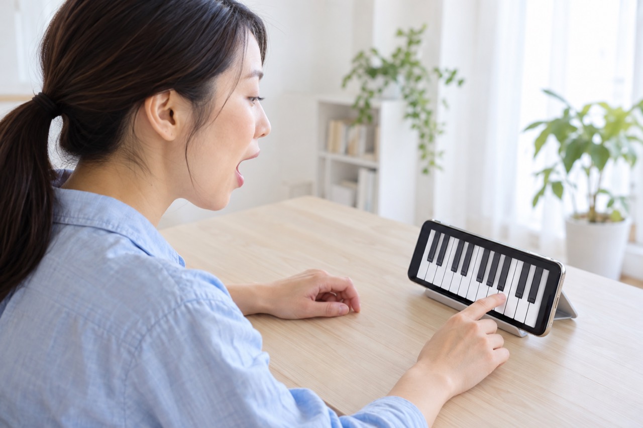 机に立てたスマホの鍵盤の画面と向かい合って、音に合わせて声を出す人